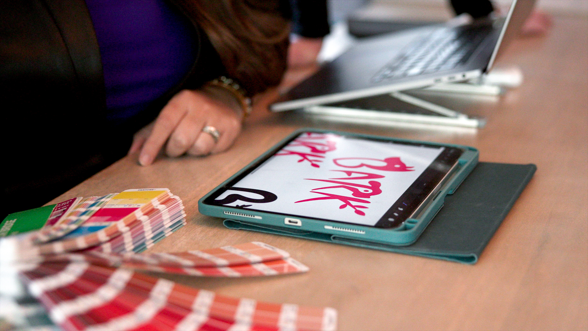 A hand sketching on an iPad with Pantone color swatches on a desk surface